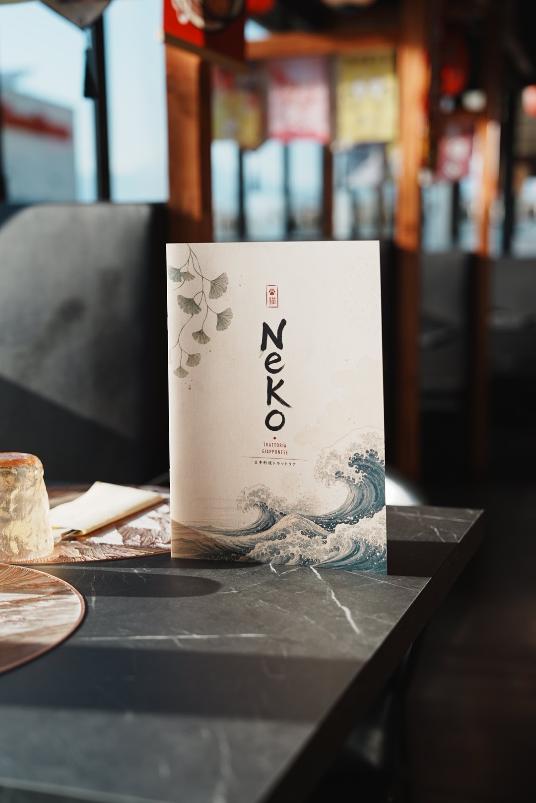 Il menu Neko fotografato nel ristorante, con carta avorio, foglie di ginkgo e un’onda illustrata.
